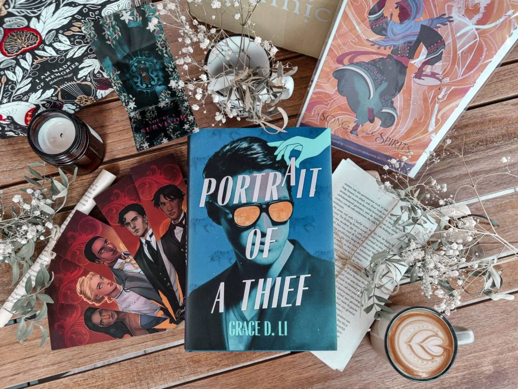Pictured here are the book 'Portait of a Thief' by Grace D. Li and a bunch of goodies by Illumicrate. They are surrounded by white flowers, a candle and a cup with coffee in it.