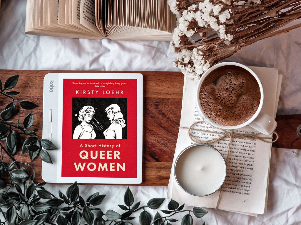 Picture of a Kobo e-reader with the cover of the book 'A Short History of Queer Women' by Kirsty Loehr on it. Surrounding it are open books, dried flowers, a plant, a candle and a mug with hot cocoa. 