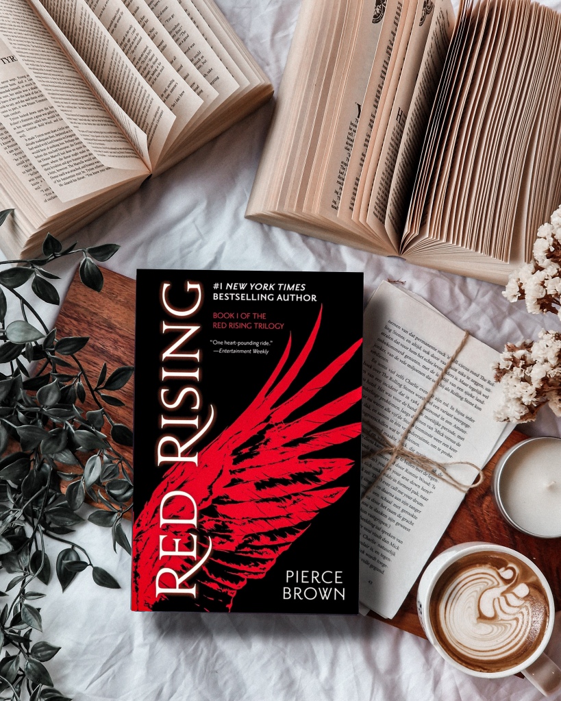 Picture of the book 'Red Rising' by Pierce Brown surounded by open books; flowers and a green plant.