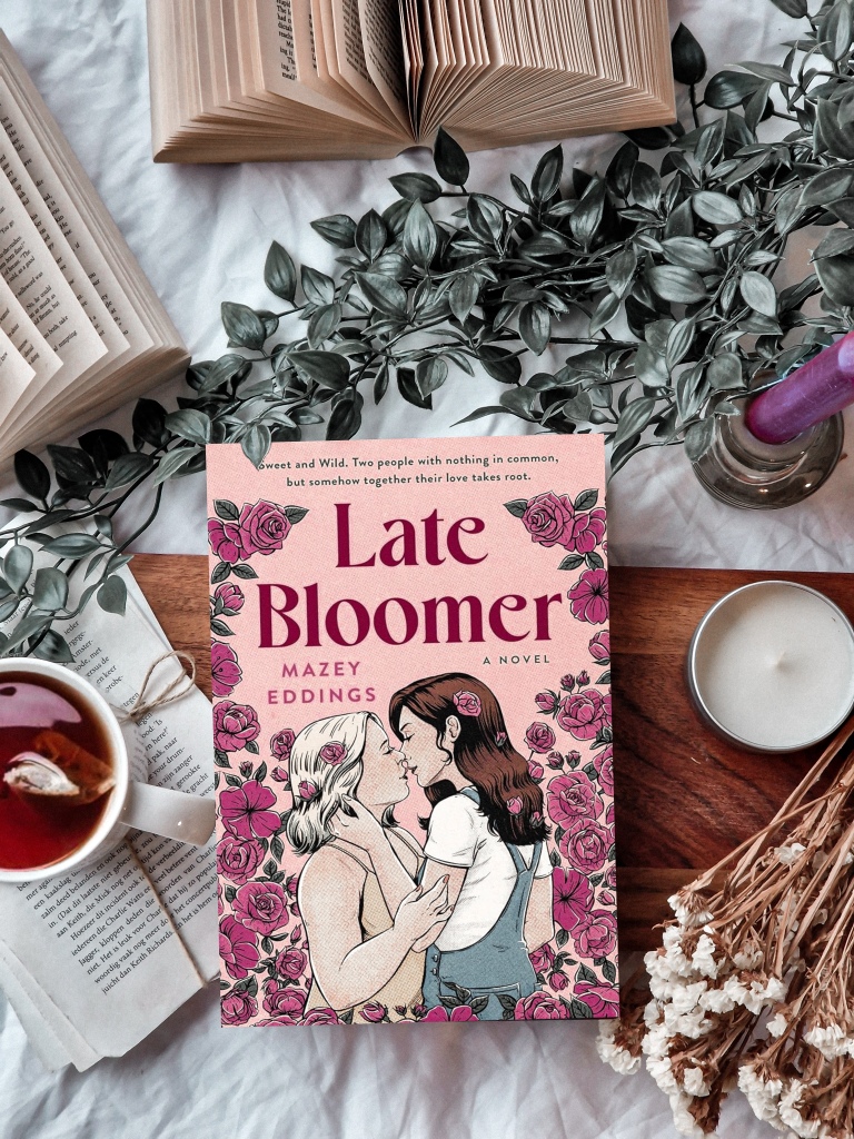 Book 'Late Bloomer' surrouned by dried flowers, green plants a candle, open books and tea.