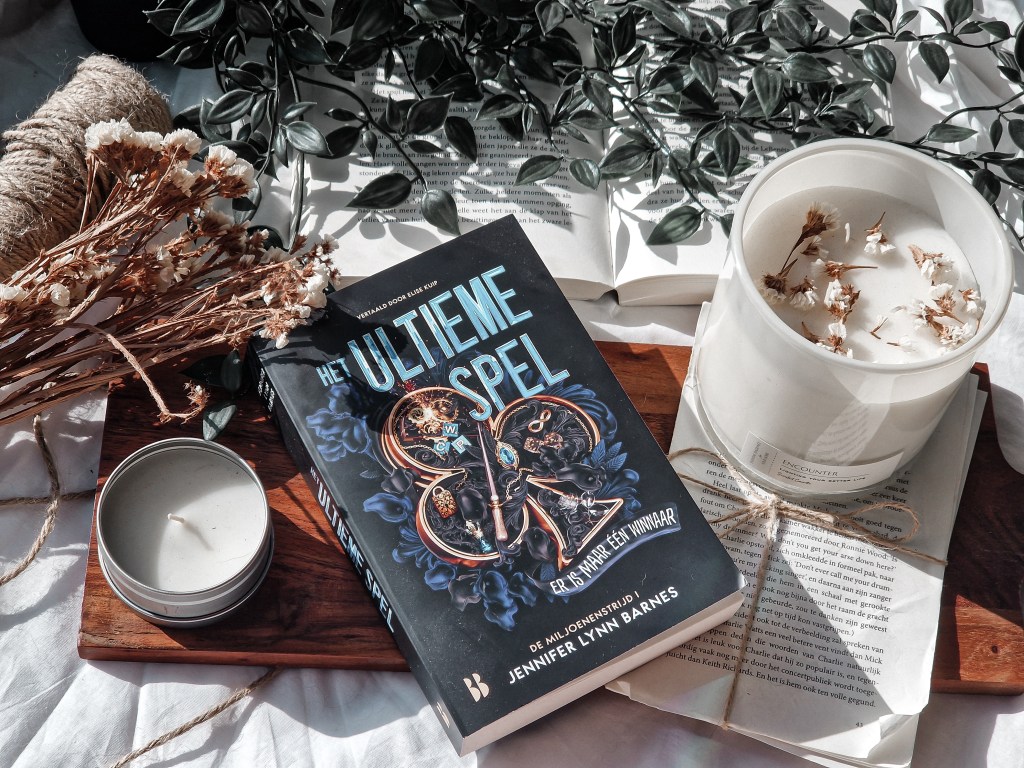 Deze foto toont het boek Het Ultieme Spel van Jennifer Lynn Barnes, stijlvol uitgestald op een houten plank. Naast het boek liggen twee kaarsen: een in een metalen blikje en een grotere witte kaars met gedroogde bloemen op de bovenkant. Links van het boek ligt een bosje gedroogde bloemen, en op de achtergrond is een open boek te zien met een blad dat gedeeltelijk is omgevouwen. Het geheel is omringd door groene bladeren, wat een serene en sfeervolle uitstraling geeft aan de compositie. De foto heeft een warme, natuurlijke lichtinval die de scène nog rustiger maakt.