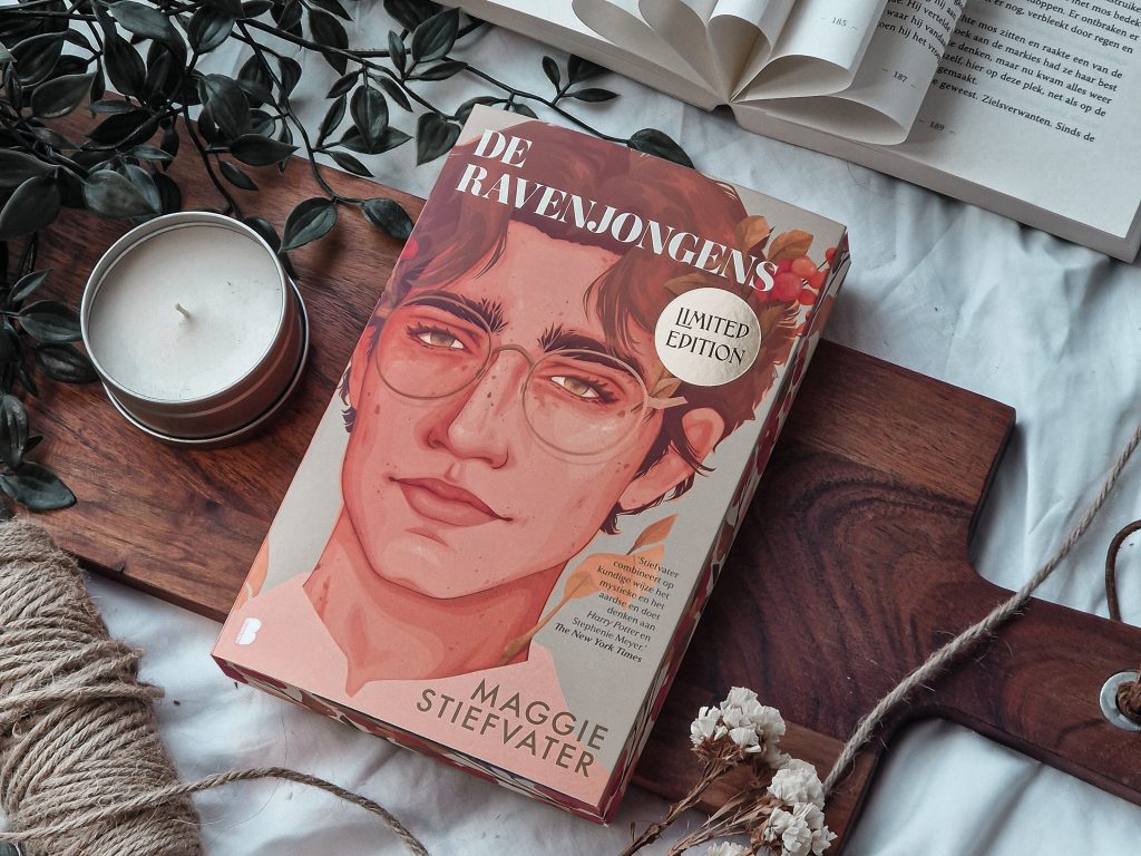 Een esthetische foto van het boek 'De Ravenjongens' door Maggie Stiefvater (limited edition), liggend op een houten plank naast een opengeslagen boek, een brandende kaars, touw, en gedroogde bloemen. De scène is omgeven door groene bladeren voor een rustieke, sfeervolle setting.