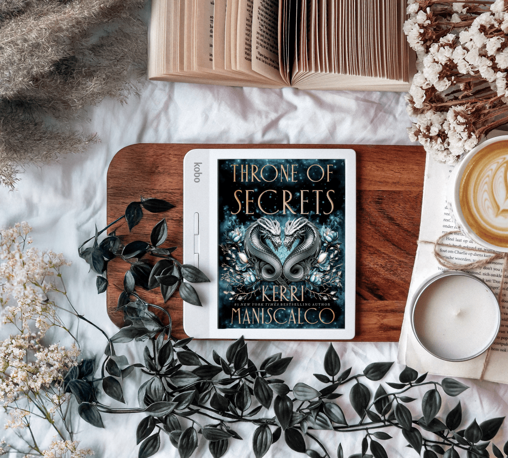A flat-lay composition featuring an e-reader displaying the book cover of Throne of Secrets by Kerri Maniscalco. The cover depicts two entwined serpents surrounded by intricate floral details. The e-reader is placed on a wooden tray, surrounded by greenery, dried flowers, an open book, and a cup of latte, creating a cozy and aesthetic bookish vibe.