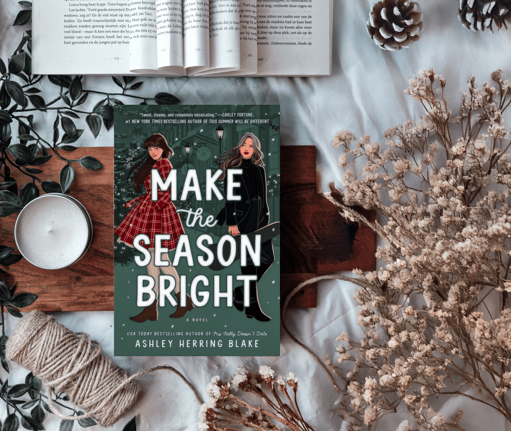 Flat lay of the book Make the Season Bright by Ashley Herring Blake, featuring a festive cover with two women in winter outfits against a background of Christmas lights and trees. The book is surrounded by cozy elements including an open book, candle, pinecones, twine, greenery, and dried flowers on a wooden and fabric backdrop.