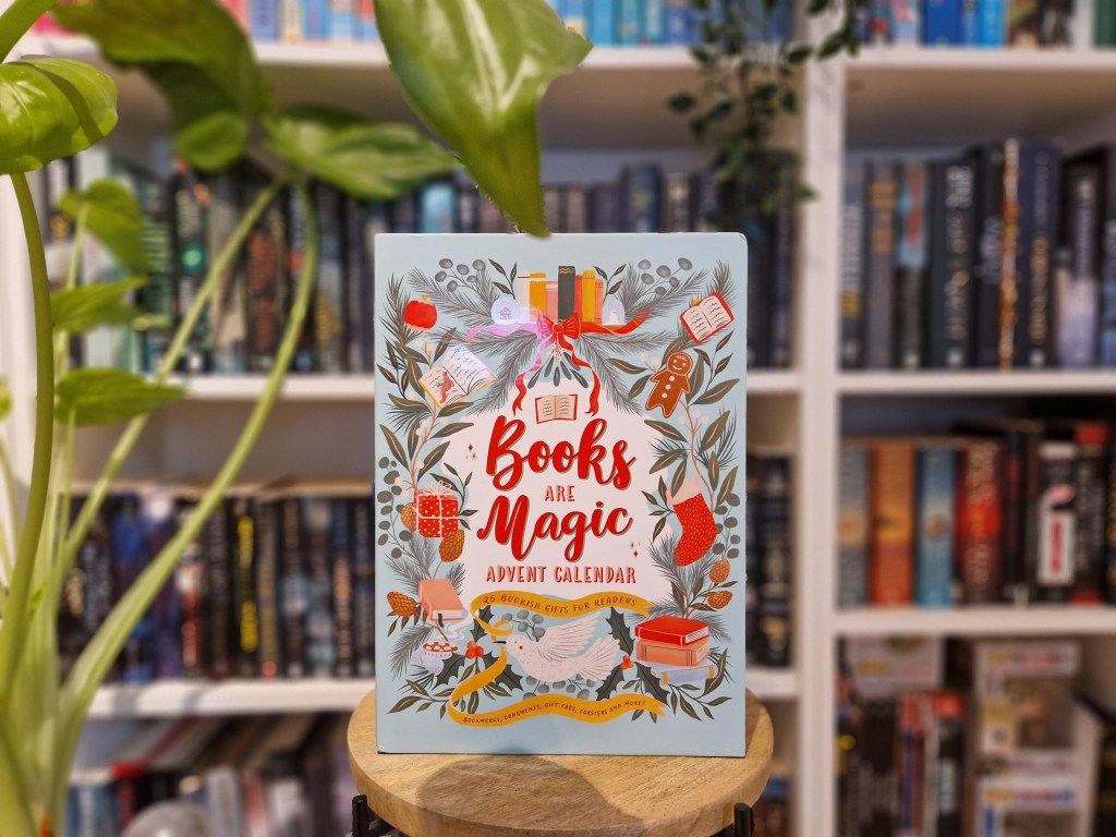 De Books Are Magic Advent Calendar in gesloten vorm, met een geïllustreerde feestelijke omslag met boeken, hulsttakken, peperkoek en decoratieve elementen. De tekst luidt: "Books Are Magic Advent Calendar: 25 Bookish Gifts for Readers." Op de achtergrond staan boekenplanken vol boeken en een groene plant voor een warme, boekige uitstraling.