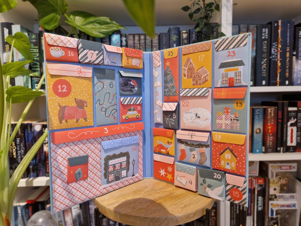 Een geopende Books Are Magic Advent Calendar met kleurrijke genummerde vakjes, elk versierd met feestelijke ontwerpen zoals een teckel in een trui, kerstverlichting, een boekwinkel en een peperkoekhuisje. Op de achtergrond staan boekenplanken vol boeken, wat een gezellige boekige sfeer creëert.