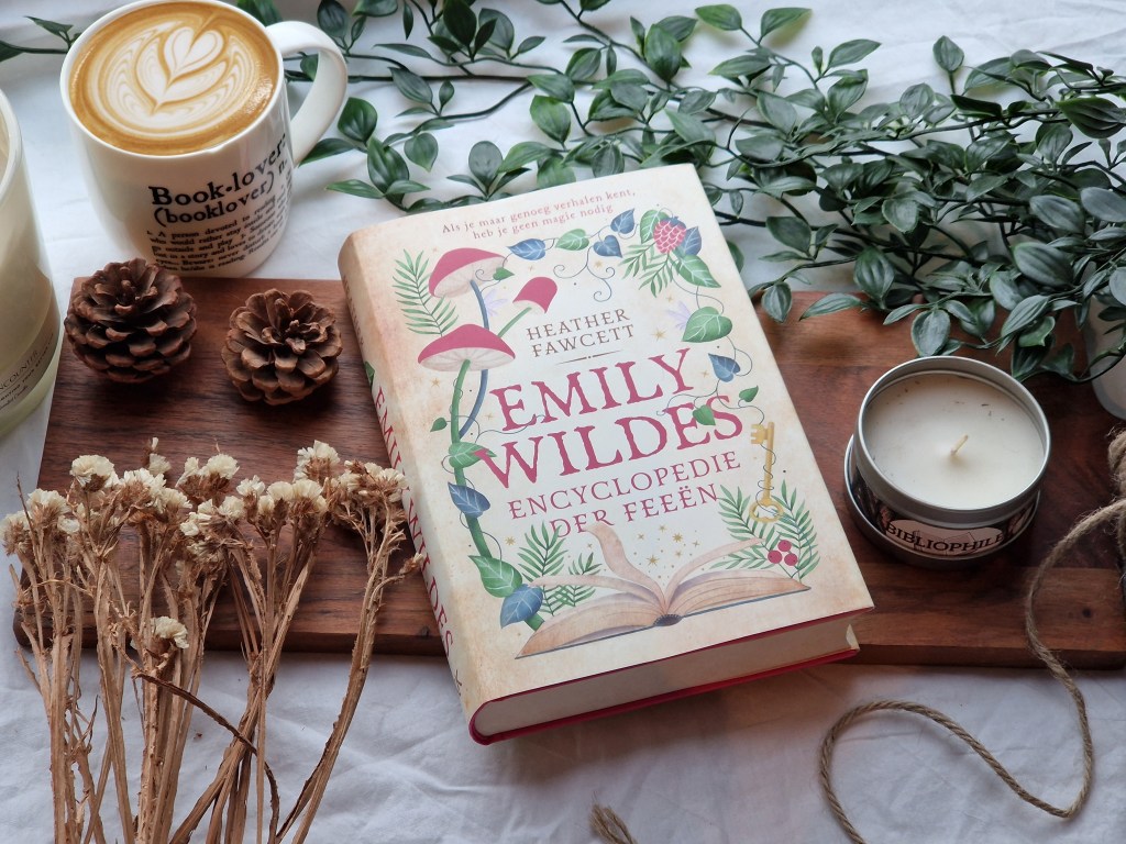 The image showcases a cozy flat-lay featuring the book Emily Wilde's Encyclopaedia of Faeries by Heather Fawcett. The book rests on a wooden board, surrounded by natural elements like pinecones, dried flowers, and a green plant. On the left, there’s a cup of coffee with latte art in a mug labeled "Book-lover (booklover)," while to the right, a lit candle and a smaller candle tin with the word "bibliophile" add to the warm ambiance. The soft lighting and earthy tones create a perfect autumnal vibe.