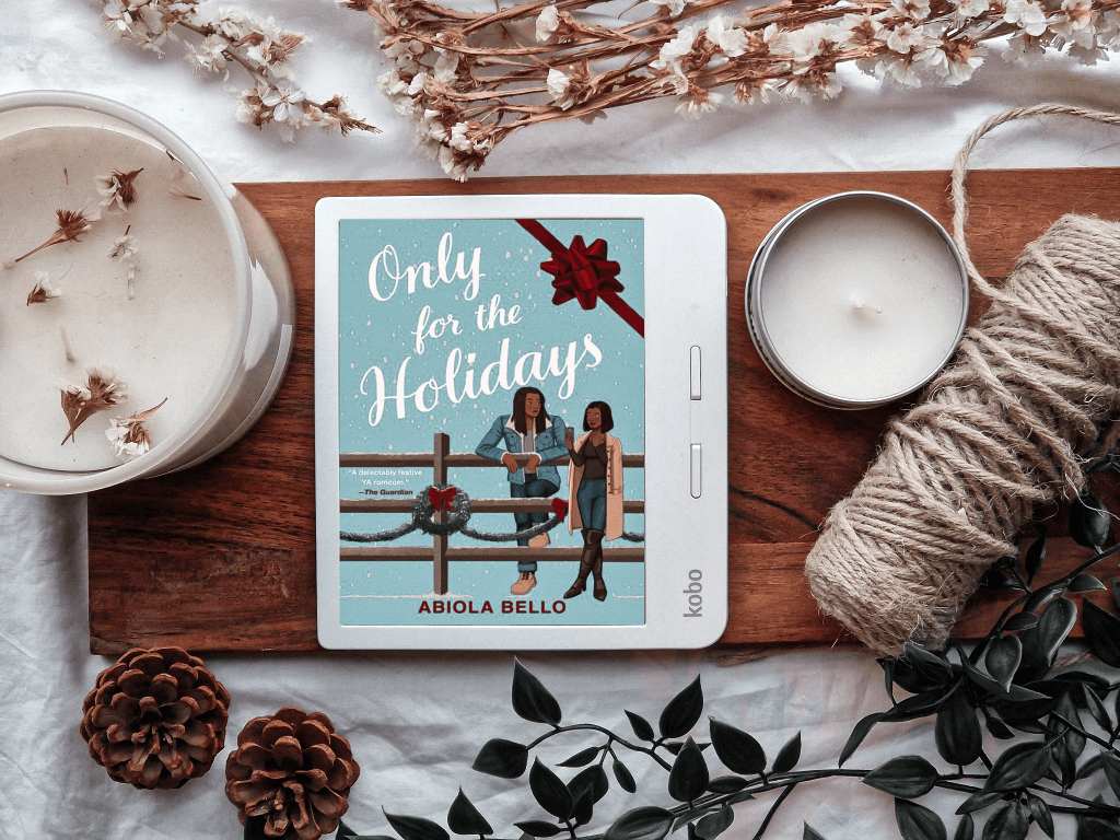 The image features a cozy and festive flat-lay showcasing a Kobo e-reader displaying the cover of the book Only for the Holidays by Abiola Bello. The cover art shows two characters standing near a wooden fence adorned with a holiday wreath, set against a snowy background. Surrounding the e-reader are aesthetic elements including candles, twine, pinecones, greenery, and dried flowers, evoking a warm and seasonal holiday atmosphere.