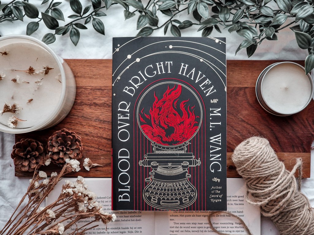 A flat lay of Blood Over Bright Haven by M.L. Wang. The book’s striking black and red cover, featuring a flaming typewriter illustration, is surrounded by cozy, natural elements like dried flowers, pinecones, candles, and a spool of twine, all set on a wooden surface with greenery in the background.
