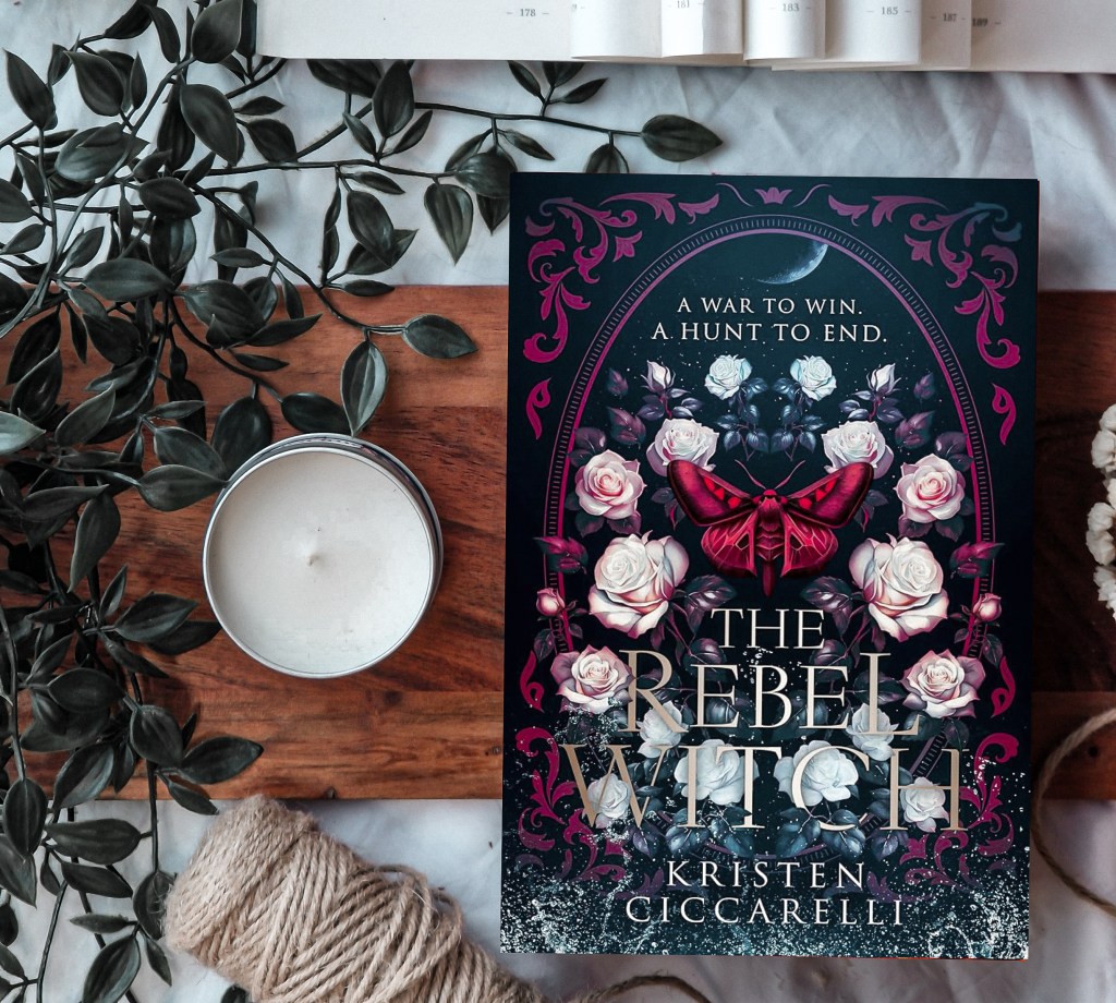 A flat-lay photograph of 'The Rebel Witch' by Kristen Ciccarelli, featuring the book with an intricate dark floral cover design. The cover showcases white and pink roses, a red moth, and the tagline "A war to win. A hunt to end." Surrounding the book are aesthetic elements, including a white candle in a tin, green trailing leaves, an open book, and a spool of twine, all arranged on a wooden surface with a cozy and atmospheric feel.
