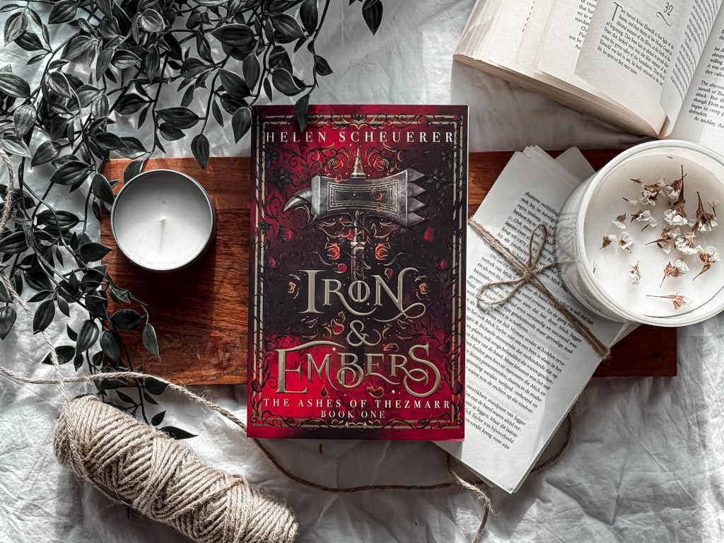 A flat lay of the fantasy novel Iron & Embers by Helen Scheuerer, surrounded by cozy elements including an open book, a lit white candle with dried flowers, a spool of twine, and leafy green vines, all arranged on a wooden tray and soft white fabric background.
