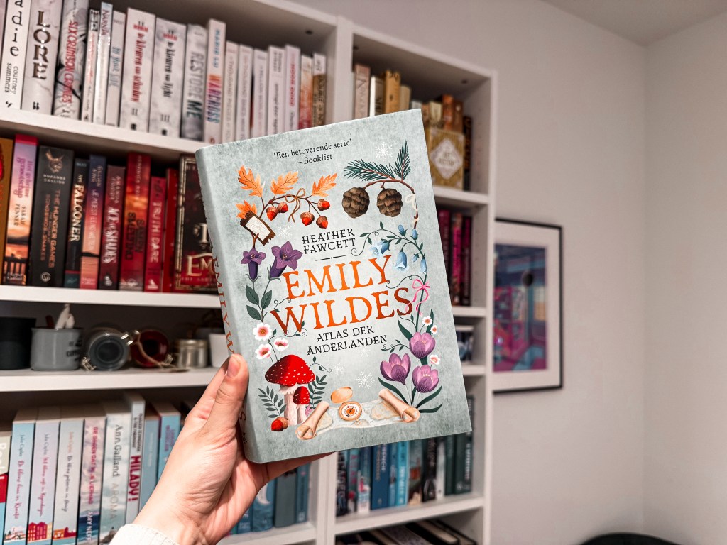 A hand holding the Dutch edition of Emily Wilde’s Map of the Otherlands by Heather Fawcett, titled Emily Wilde’s Atlas der Anderlanden. The book features a botanical cover design with mushrooms, pinecones, and flowers. In the background is a cozy bookshelf filled with colorful books.