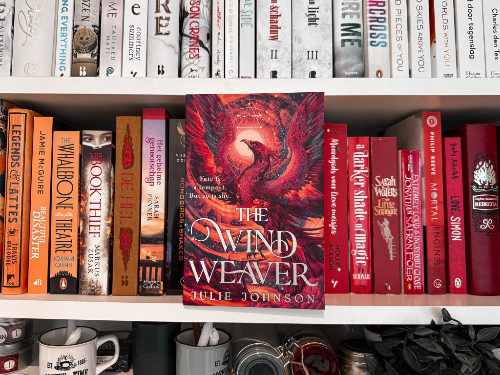 A vibrant photo of the book The Wind Weaver by Julie Johnson placed on a bookshelf, centered among other red-toned books. The book cover features a fiery red phoenix with glowing wings set against a magical, starry background. The tagline reads, "Fate is a tempest. But so is she." Surrounding books include A Darker Shade of Magic, Love, Simon, Mortal Engines, and The Book Thief. The shelf is color-organized, transitioning from orange to red books, with mugs and candles placed on the shelf below.