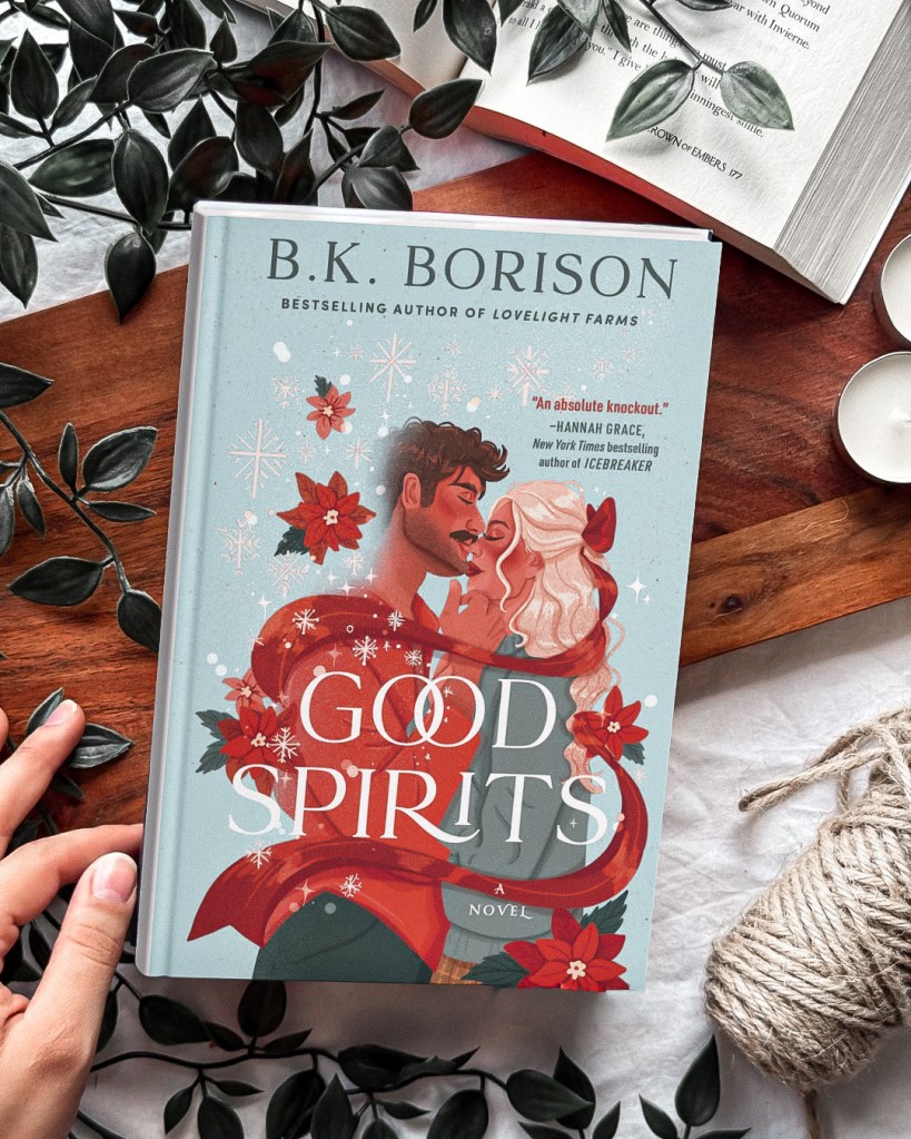 Flat lay of Good Spirits by B.K. Borison, a holiday romance novel with illustrated couple on the cover, surrounded by Christmas decor, greenery, and cozy winter elements. Perfect festive book for fans of spicy Christmas romance reads.