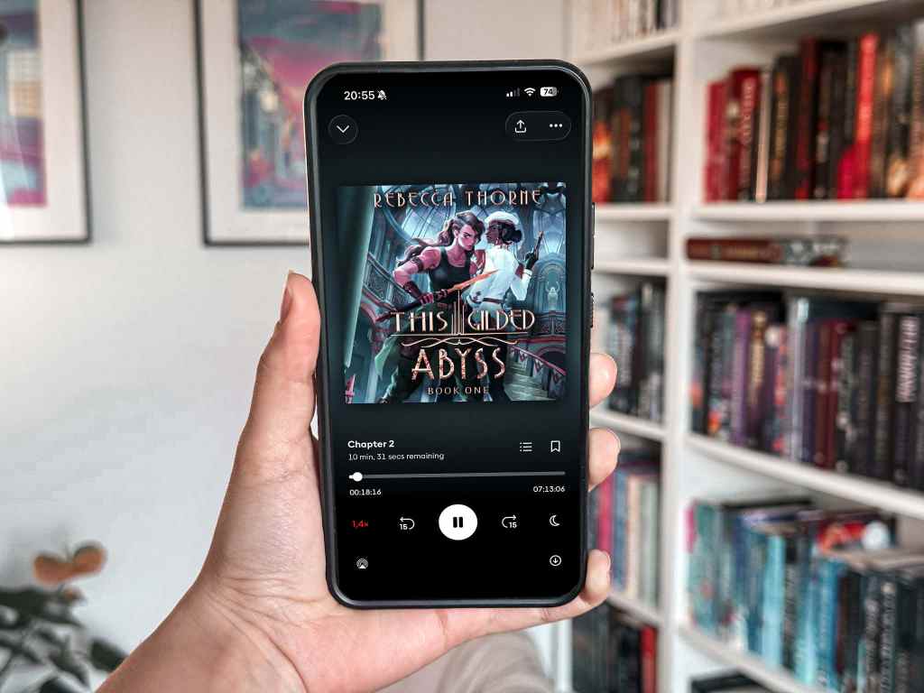 A hand holds up a smartphone displaying the audiobook This Gilded Abyss by Rebecca Thorne. The cover art shows two women standing back to back, one in a white uniform with a sword, the other in pink with glowing hands, set against a richly detailed underwater backdrop. Behind the phone is a white bookshelf filled with colorful fantasy books and framed art on the wall.