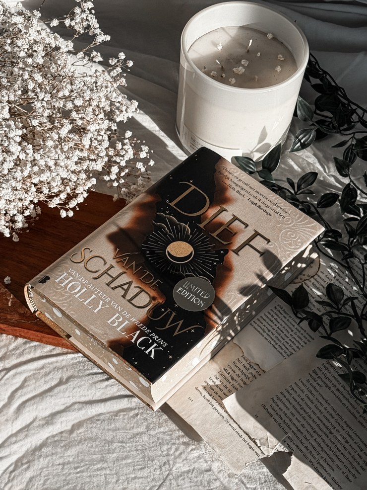 A cozy flat lay of Thief of Night (Dief van de schaduw in Dutch) by Holly Black in a limited edition hardcover, resting on soft white bedding beside a large white candle, delicate dried baby’s breath flowers, scattered book pages, and trailing dark green leaves in warm natural sunlight.